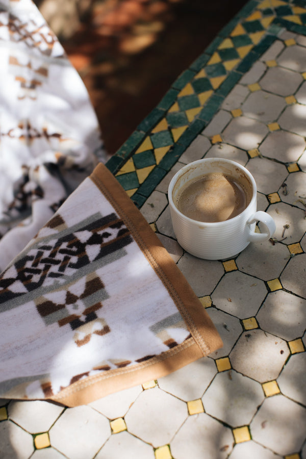Corner of the white and brown celtic blanket of mother and child on a outdoor table close up next to a coffee cup with sun spots hitting the surfaces from above