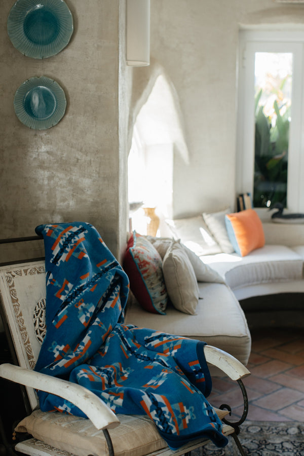 Blue parent and child blanket on a white rocking chair, on the wall behind there are teal plates and windows behind with oval walls and seating underneath